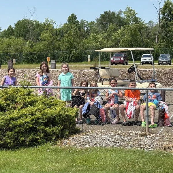 Group of children beside a golf cart near a fence and greenery.